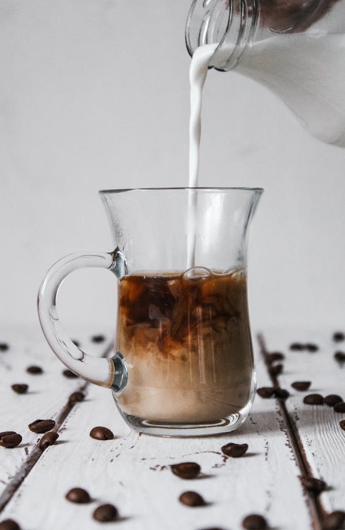 Glass mug of coffee with milk being poured, surrounded by scattered coffee beans on a wooden table.